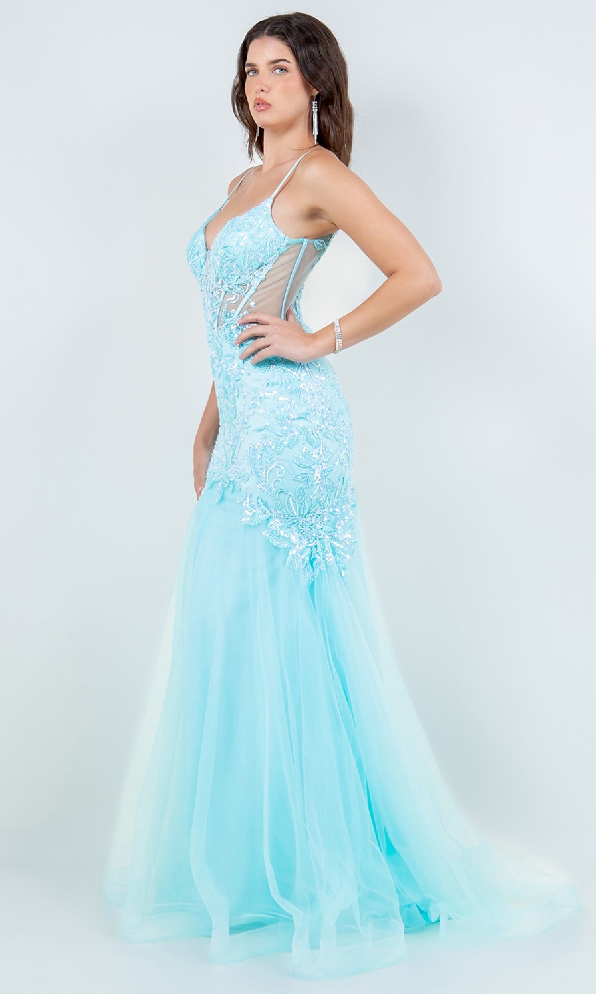 A woman stands in profile against a plain background, wearing the Cinderella Couture 8276J—a light blue, fitted sequin mermaid prom dress with sheer sides, intricate embellishments, and thin straps.